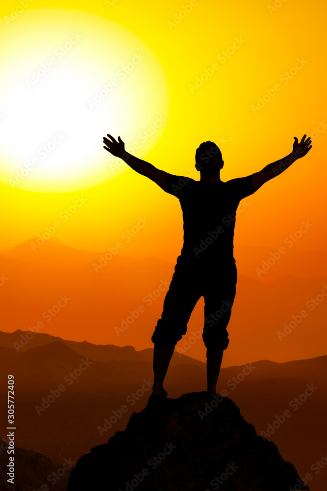 Happy celebrating winning success man at sunset or sunrise standing elated with arms raised up above her head in celebration of having reached mountain top summit goal during hiking travel trek.
