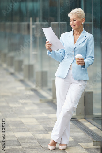 Mature businesswoman in beautiful suit reading business report and drinking coffee before meeting while standing outdoors