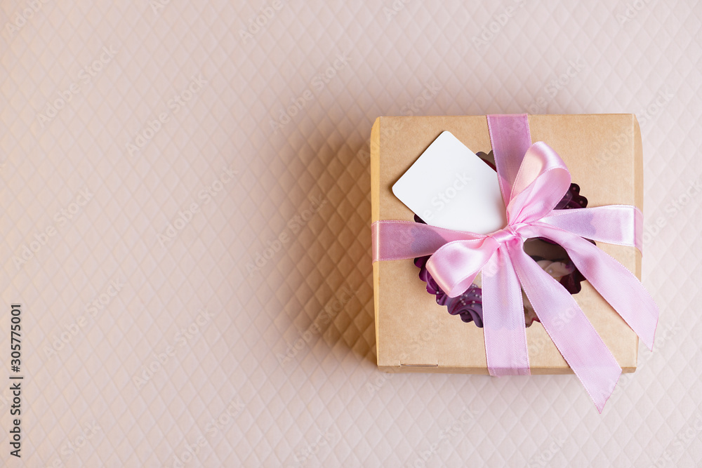 A square gift box with a pink ribbon on a light pink background. Cupcakes inside. Space for text, empty note tied over. A square gift box with a pink ribbon on a light pink background. Cupcakes inside. Space for text, empty note tied over.