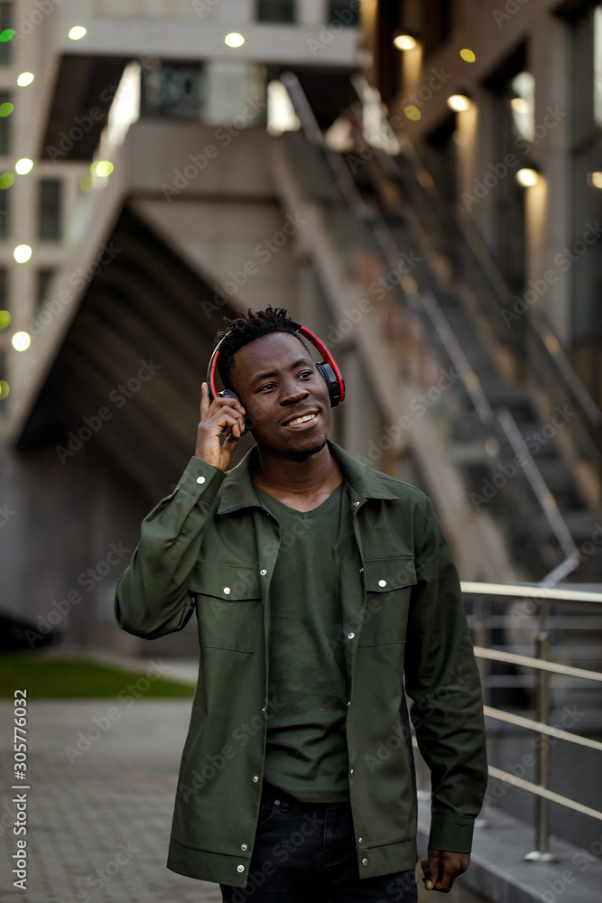 Fototapeta premium african-american man in stylish jacket in wireless headphones listening music on the street of the evening city