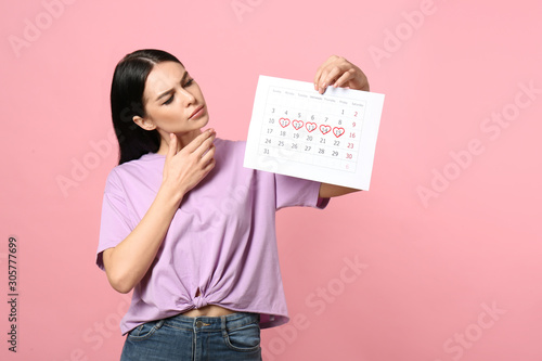 Pensive young woman holding calendar with marked menstrual cycle days on pink background photo