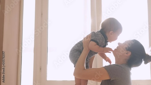 Mother and her baby son having fun and playing at home. Little kid 1 years old play with his mom arms at home near a big window, slow motion. They feel happy, smile and laugh
