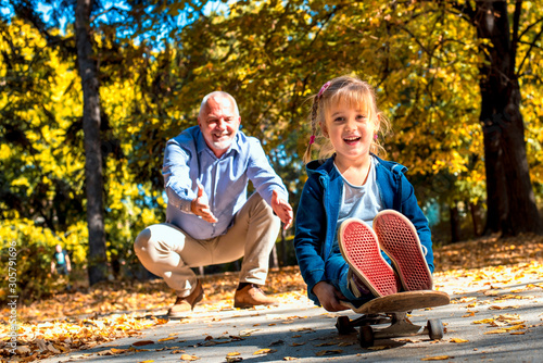 Granddaughter enjoying day with grandfather in the autumn park while riding skateboard photo