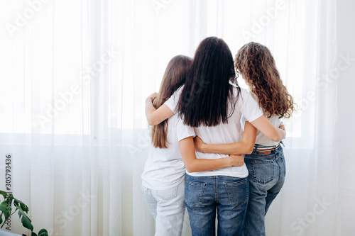 Back view of beautiful brunette woman and her daughters hugging and looking through the window, sitting at home © speed300