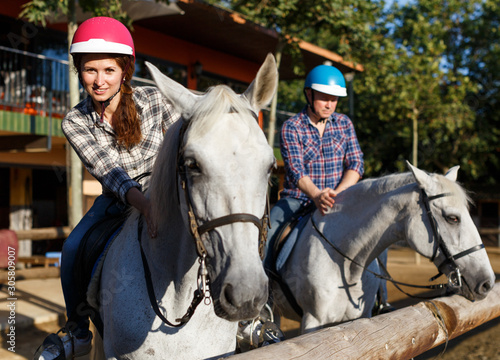 Mature couple in helmets riding by horse at barn at summer day