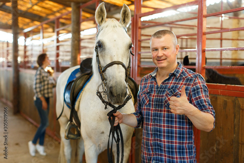 Mature positive man farmer standing with white horse at stabling