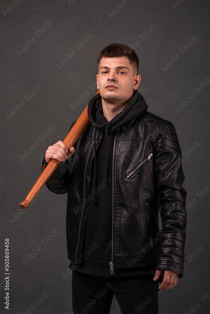 The young gue wearing leather jacket holding baseball bat in his hand at a grey wall The young gue wearing leather jacket holding baseball bat in his hand at a grey wall