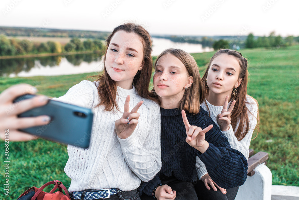 Fototapeta premium Three teenage girls, teenagers in summer city have rest after school college, hands smartphone, selfie picture video call online application on social networks. Happy smiling. Casual clothes, sweater.