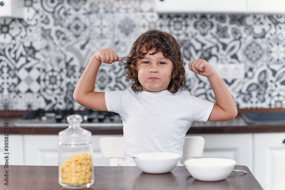 Happy little boy showing biceps muscles while eating healthy breakfast of milk and cornflakes at the stylish kitchen in the morning. Happy little boy showing biceps muscles while eating healthy breakfast of milk and cornflakes at the stylish kitchen in the morning.
