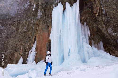 Woman hiker next to a tall frozen waterfall during a Winter hike at Seven Ladders Canyon (7 Scari) in Piatra Mare massif, Brasov county, Romania.