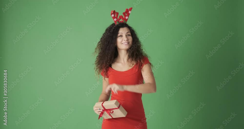 Beautiful woman in reindeer hairband holding gift while dancing and celebrating Christmas over green background Beautiful woman in reindeer hairband holding gift while dancing and celebrating Christmas over green background