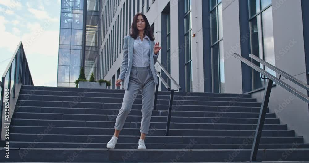 Joyful young Caucasian businesswoman stepping down the steps outside the big urban building and dancing funny as having good news or successful contract. Joyful young Caucasian businesswoman stepping down the steps outside the big urban building and dancing funny as having good news or successful contract.