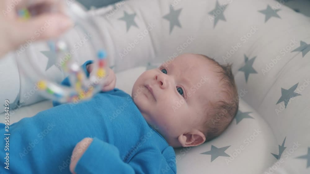 happy little newborn baby bothers arms looking at funny rattle in mother hand in cocoon with grey stars happy little newborn baby bothers arms looking at funny rattle in mother hand in cocoon with grey stars