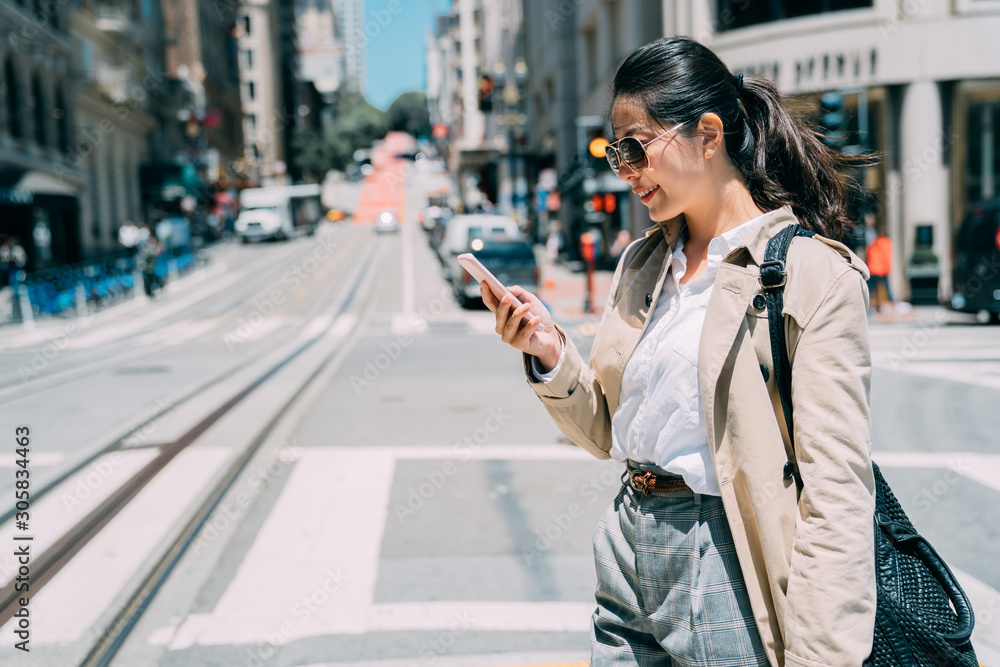 Fototapeta premium Young asian chinese businesswoman on sunny busy city street in san francisco america. woman manager texting on cellphone walking on road in urban. smiling office lady in sunglasses use mobile phone