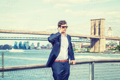 Young American Man traveling, relaxing in New York City, wearing blue suit, white shirt, sunglasses, standing by East River, talking on cell phone. Manhattan, Brooklyn bridges, boats on background..