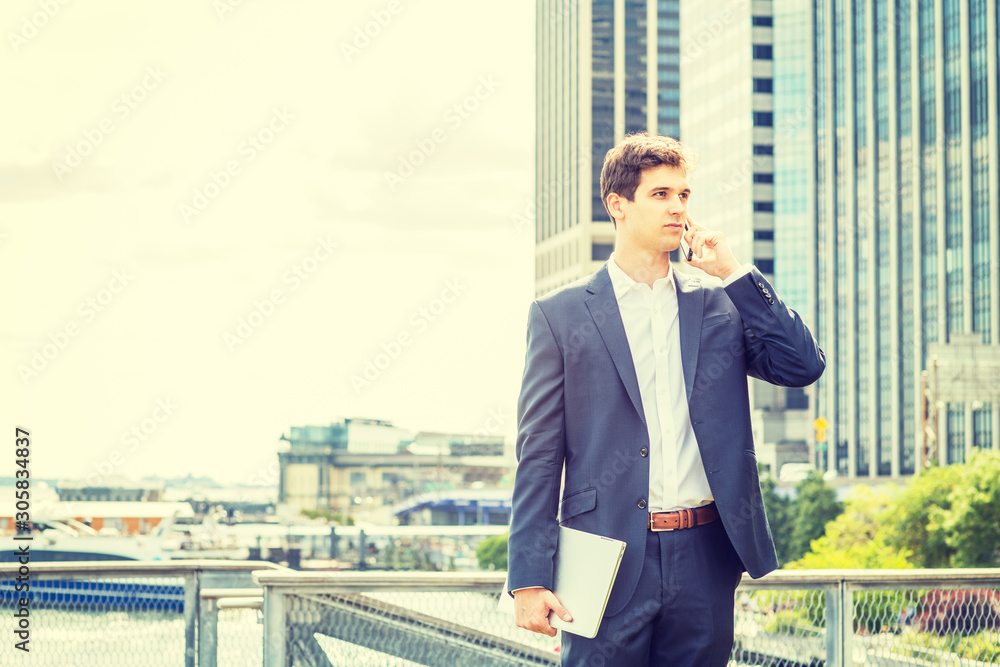Young handsome businessman traveling, working in New York City, wearing blue suit, white shirt, holding laptop computer, standing in front of business district, talking on cell phone, looking forward. Young handsome businessman traveling, working in New York City, wearing blue suit, white shirt, holding laptop computer, standing in front of business district, talking on cell phone, looking forward.