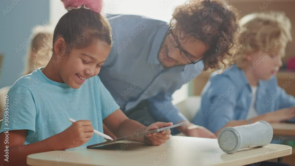 Elementary School Computer Science Class: Smart Girl Uses Digital Tablet Computer, Friendly Teacher Helps Her by Explaining Lesson and Asignment. Children Getting Modern Education Elementary School Computer Science Class: Smart Girl Uses Digital Tablet Computer, Friendly Teacher Helps Her by Explaining Lesson and Asignment. Children Getting Modern Education