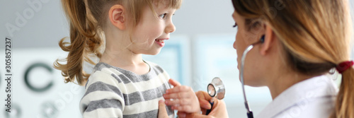 Portrait of cheerful child covering chest by hands. Smiling baby in striped sweater visiting family doctor. Health care and prevention concept. Blurred background