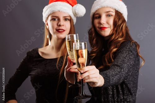 Two attractive cheerful girls with curly long hair in Christmas hat smiling, holding a champagne, posing to the camera, isolated on gray background