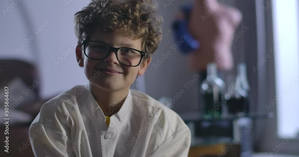 Portrait of smart Caucasian boy in eyeglasses looking at camera and smiling. Close-up of intelligent child with curly hair sitting at the background of flasks with chemicals. Cinema 4k ProRes HQ. Portrait of smart Caucasian boy in eyeglasses looking at camera and smiling. Close-up of intelligent child with curly hair sitting at the background of flasks with chemicals. Cinema 4k ProRes HQ.