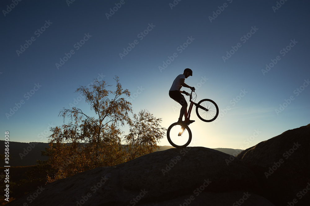 Silhouette of athlete cyclist standing on back wheel on trial bicycle. Sportsman rider balancing on the edge of big boulder on the top of mountain at sunset. Concept of extreme sport active lifestyle Silhouette of athlete cyclist standing on back wheel on trial bicycle. Sportsman rider balancing on the edge of big boulder on the top of mountain at sunset. Concept of extreme sport active lifestyle