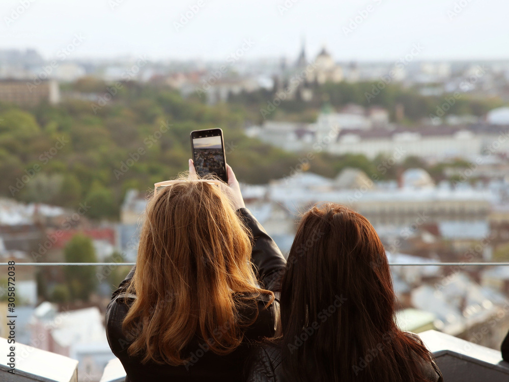 Two girls take a panorama photo of the ancient city with a smartphone. An observation deck at the town hall of the central Rinok square of the city of Lviv in Ukraine. Tourist selfie place on sunset Two girls take a panorama photo of the ancient city with a smartphone. An observation deck at the town hall of the central Rinok square of the city of Lviv in Ukraine. Tourist selfie place on sunset