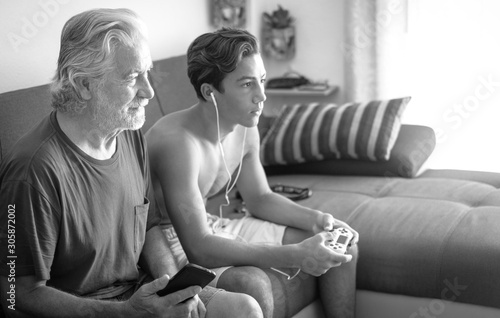 Handsome male teenager plays video games while his senior grandfather follows the game. Different generations and the same concept of interest. Love and emotion. Sitting indoors on a brown couch