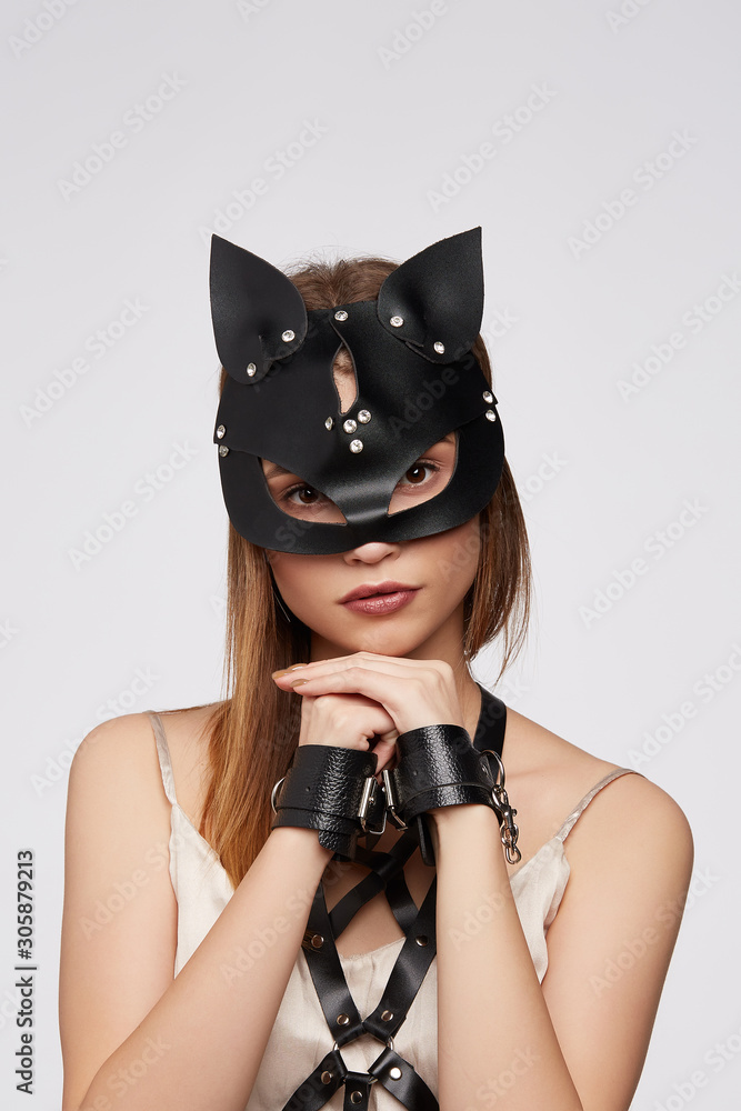 Fotka „Medium close-up shot of a pretty lady in a white undervest, a black  leather harness made of straps with rivets, leather cuffs and a black  leather catwoman mask with rhinestones and