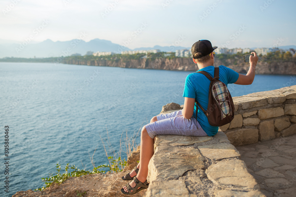 Young kid taking selfie using modern mobile smartphone posing outdoor at sunny scenic seascape. Happy travel and modern technology concept. Horiozntal color photography.