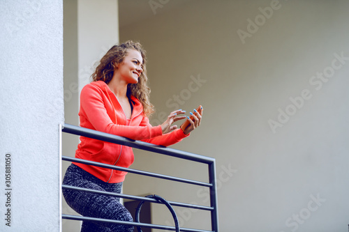 Smiling beautiful caucasian woman in sportswear and with curly hair leaning on railing and using smart phone.