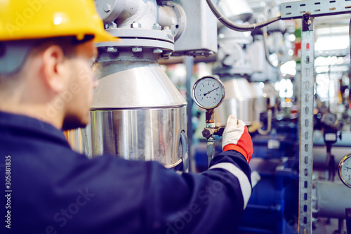 Rear view of hardworking blue collar worker in protective working suit and with helmet adjusting temperature on boiler while standing in factory. photo