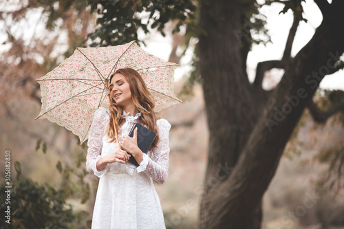 Happy young woman 24-26 year old holding paper book and vintage small umbrella walking in park outdoors.