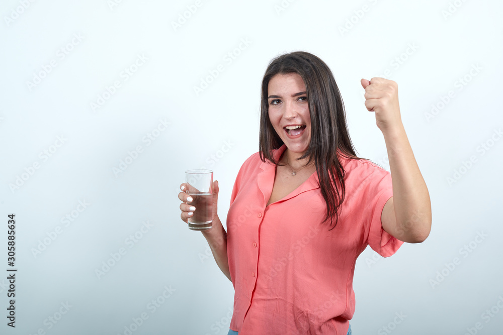 Pretty caucasian young woman keeping glass of water, doing winner gesture with fist isolated on white background in studio in pink shirt. People sincere emotions, lifestyle concept. Pretty caucasian young woman keeping glass of water, doing winner gesture with fist isolated on white background in studio in pink shirt. People sincere emotions, lifestyle concept.