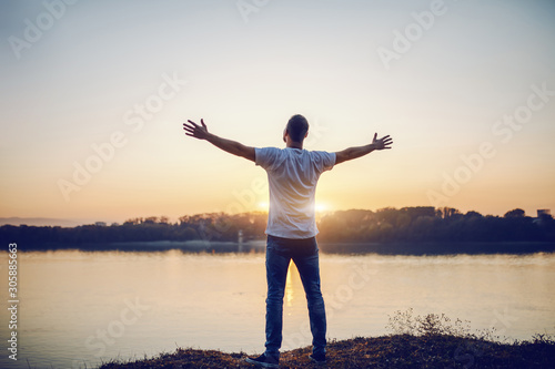 Fotografija Rear view of handsome caucasian man standing with opened arms on cliff and looking at river and beautiful sunset