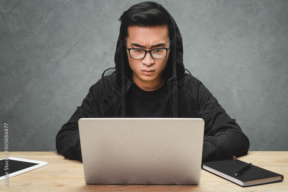 asian hacker in glasses using laptop and sitting at table asian hacker in glasses using laptop and sitting at table