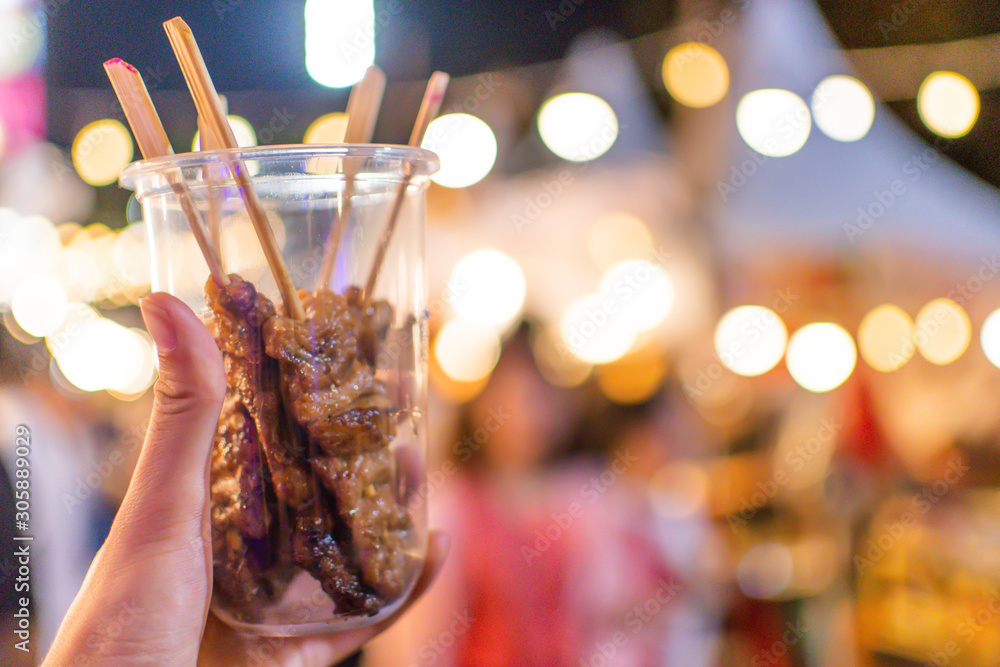 A hand holding a cup of beef grill. With the bokeh backgrounds of blur people and market.