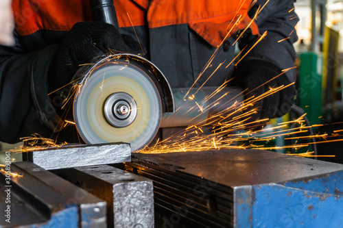 The worker cuts the metal with an electric angle grinder, sparks fly around.