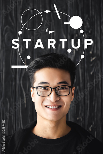 smiling asian man in glasses looking at camera on wooden background with startup illustration photo