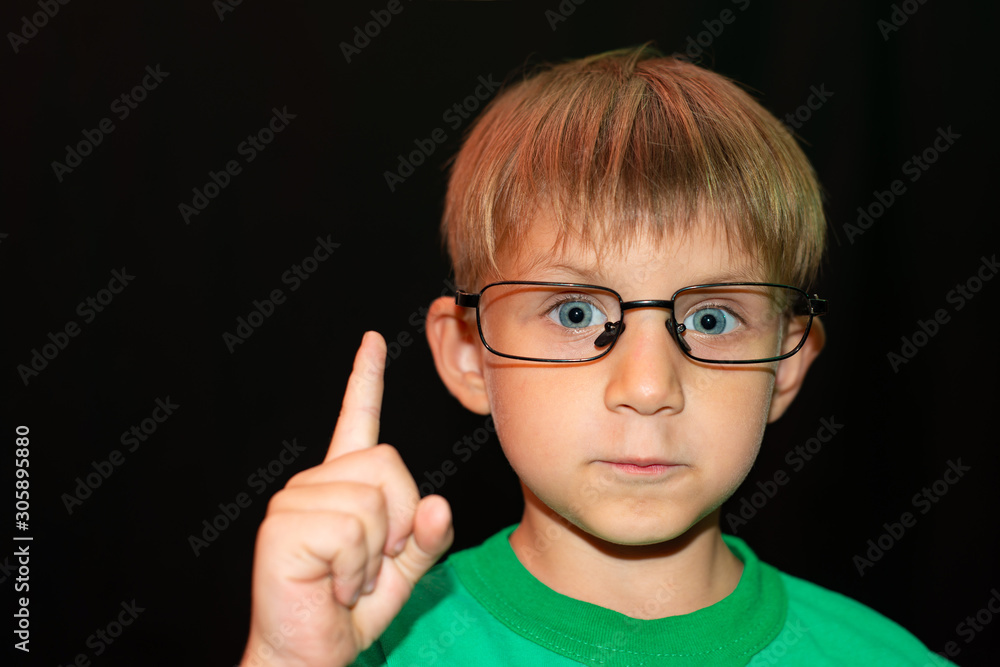 The boy in glasses on a black background looks at the camera and shows a finger to the top.