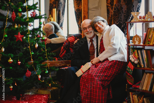 An elegant old couple is sitting by the Christmas tree. Grandparents give each other gifts.