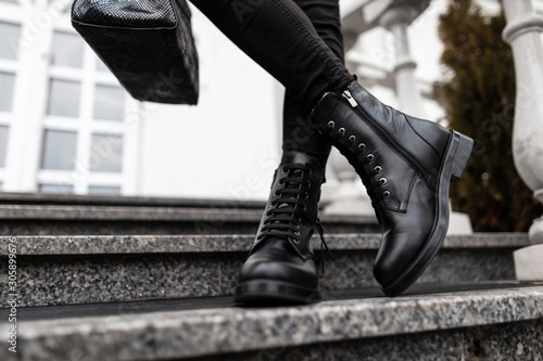 Young trendy woman in stylish leather black boots in jeans with a bag stands on the stone steps in the city. Close up of female legs in fashion footwear. New collection of shoes and accessories.