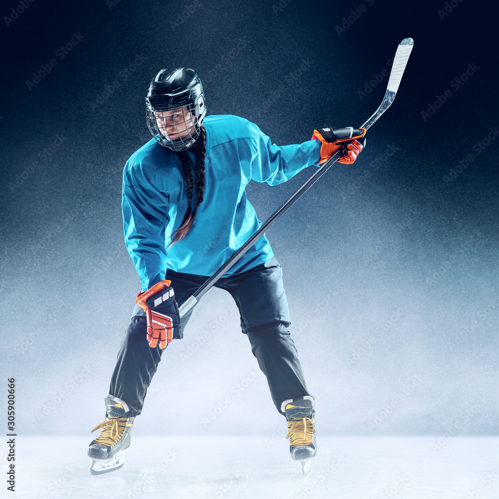 Obraz premium Young female hockey player with the stick on ice court and blue background. Sportswoman wearing equipment and helmet practicing. Concept of sport, healthy lifestyle, motion, movement, action.