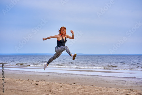 A middle aged woman with red hair jumps while jogging. A woman practices gymnastics on the sandy bank of a large river. Cloudy spring morning.