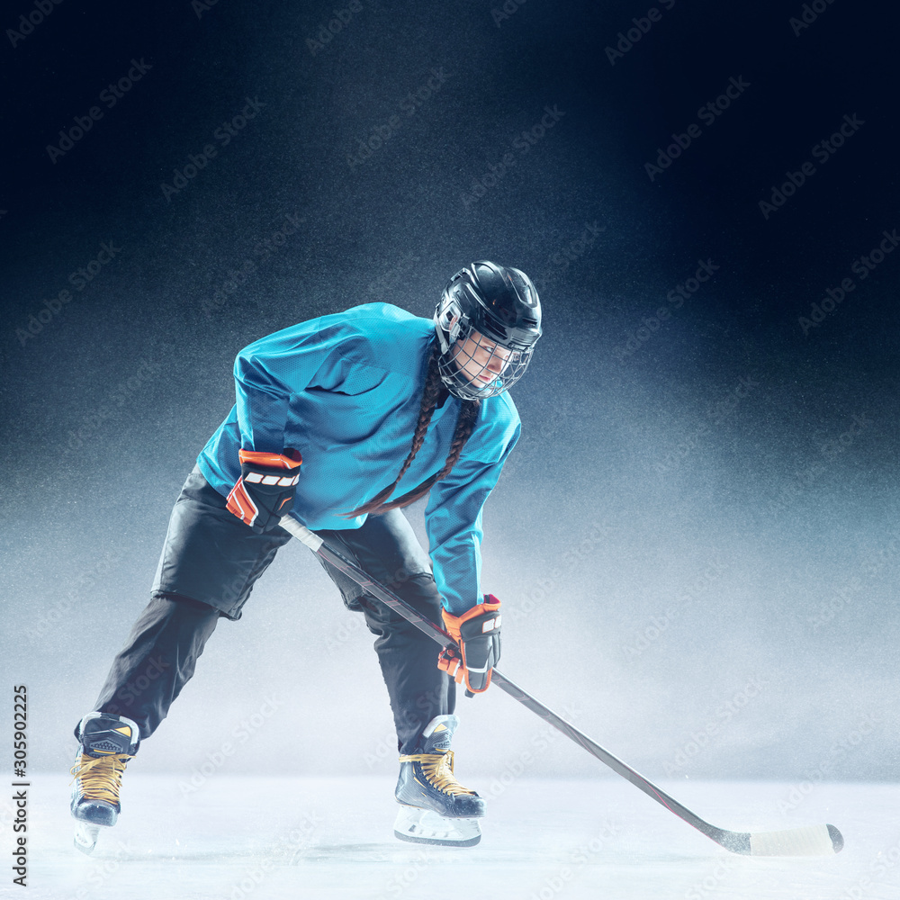 Obraz premium Young female hockey player with the stick on ice court and blue background. Sportswoman wearing equipment and helmet practicing. Concept of sport, healthy lifestyle, motion, movement, action.