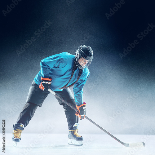 Young female hockey player with the stick on ice court and blue background. Sportswoman wearing equipment and helmet practicing. Concept of sport, healthy lifestyle, motion, movement, action.