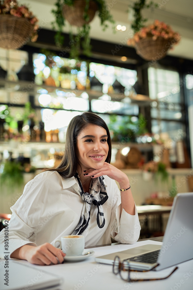 Attractive caucasian elegant brunette sitting in cafe, holdingcoffee and looking at away. On table is laptop. Attractive caucasian elegant brunette sitting in cafe, holdingcoffee and looking at away. On table is laptop.