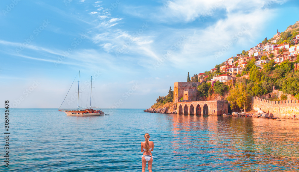 Obraz premium Beautiful young woman in white bikini stands on the sea - Landscape of ancient shipyard near of Kizil Kule tower - Alanya peninsula, Turkey