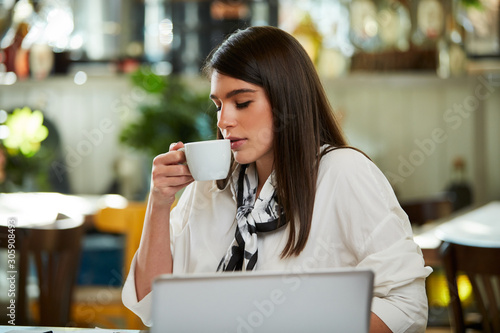 Young caucasian businesswoman sitting in cafe, taking a break from work and drinking fresh coffee. On table are laptop and paperwork.
