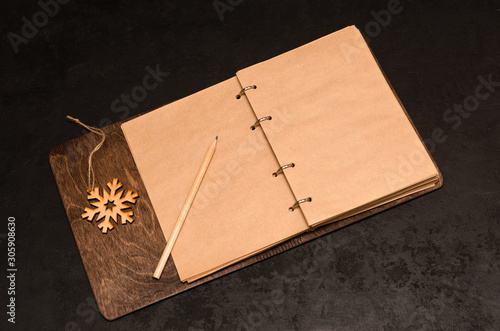 Kraft notebook with blank sheets in a wooden cover with space for your text, pencil and wooden snowflake on black background. Plan next year concept. To do list. Christmas background. Mockup