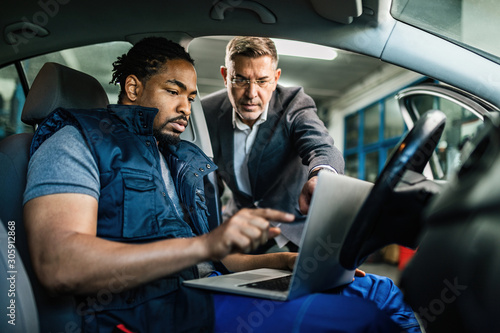 African American auto mechanic and his customer cooperating while using laptop in repair shop.